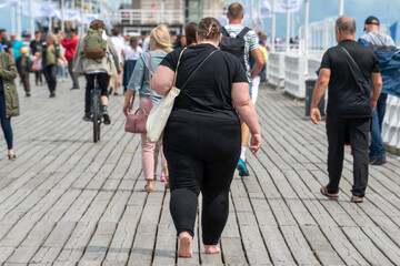 Overweight woman walking on the pier among people with normal weight. Barefoot obese woman wearing...