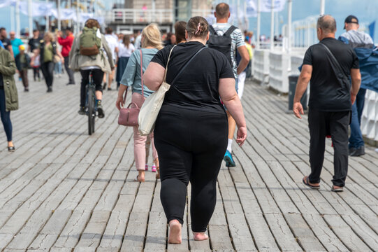 Overweight woman walking on the pier among people with normal weight. Barefoot obese woman wearing black. Obesity, losing weight.