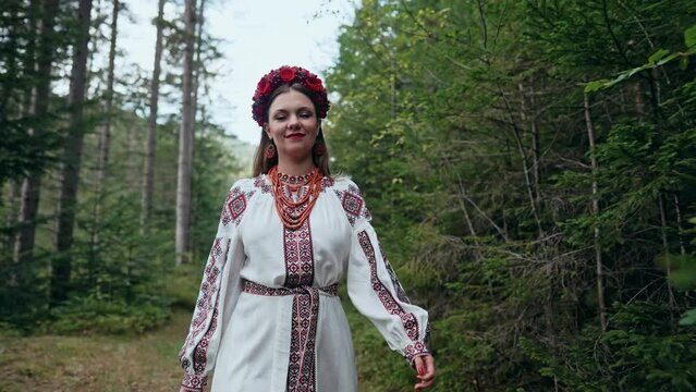 Ukrainian woman walking in fir forest, Carpathian mountains nature. Girl in traditional embroidery vyshyvanka dress, red wreath, ancient jewelry. Ukraine, freedom, ethnic national costume