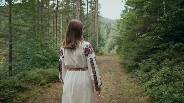Back view of unrecognizable ukrainian woman walking in fir forest, Carpathian mountains nature. Girl in traditional embroidery vyshyvanka dress. Ukraine, freedom, ethnic national costume