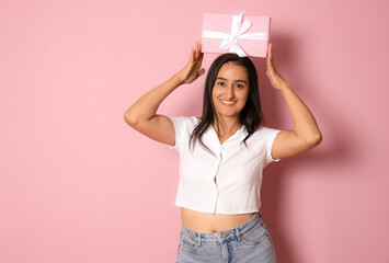 Portrait of an excited hispanic happy girl holding a gift box present on her head and looking at camera isolated over pink background