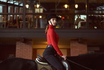 Young beautiful woman sitting horseback in dark indoor manege  and looking at the camera. Woman...