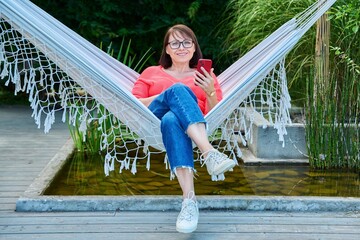 Middle aged woman with smartphone sitting in hammock