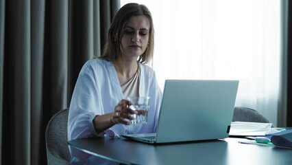 Focused concentrated female working or studying, drinking water and looking on laptop screen.