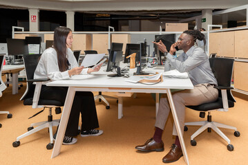 two multi-ethnic business partners sitting in front of each other at a table with computers working together on a project in a coworking space