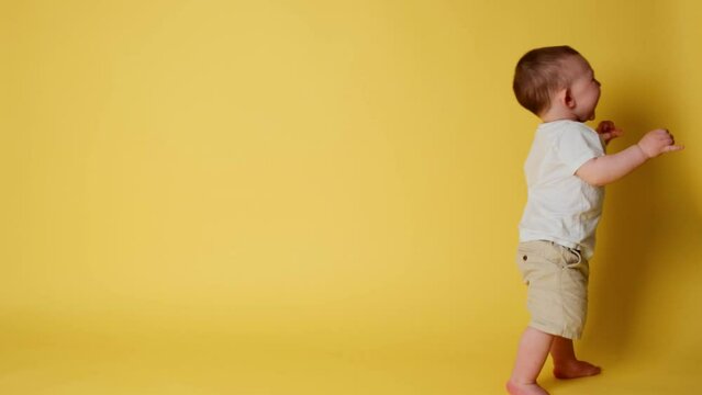 Happy toddler baby learns to walk and takes his first steps. Child boy walks for the first time on a studio yellow background. Kid age one year