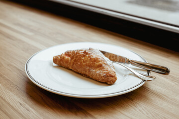 A croissant on a white plate, cutlery on a wooden window-will