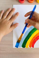 A child's hand draws a rainbow with a brush.Many cans of red yellow green color on the mirror table in the classroom.Female fingers hold open blue paint.The concept of school art education.