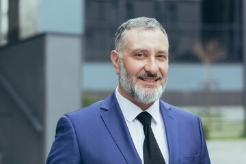 Portrait of senior handsome businessman man with gray hair and brown hair in business suit with tie. Near the office center, looks at the camera, smiles. - Powered by Adobe