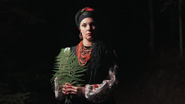 Authentic woman in traditional ukrainian handkerchief, necklace and costume with fern in forest at night. Lady in national dress - vyshyvanka, ancient coral beads. Concept of witch.