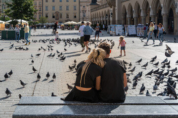 Obraz premium Couple sitting on a marble bench outdoors in the city square on a sunny summer day, traditional young couple, tourists in Krakow, close relationship, love