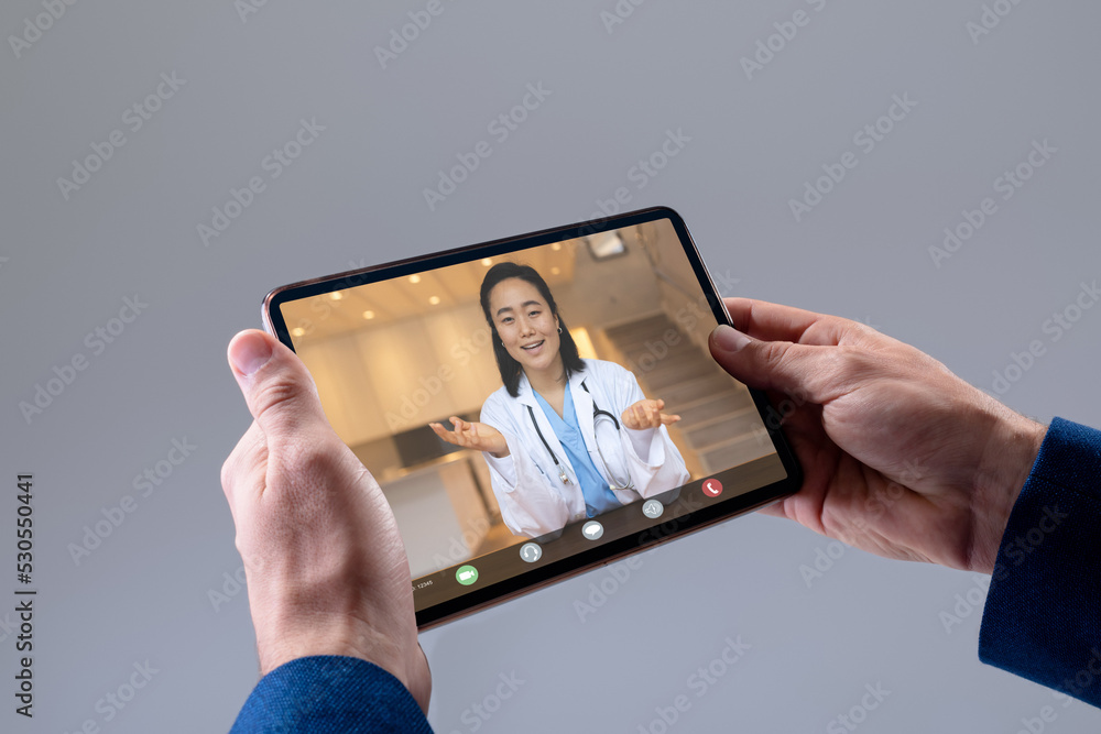 Wall mural Hands of caucasian businessman making video call on tablet with asian female doctor on screen