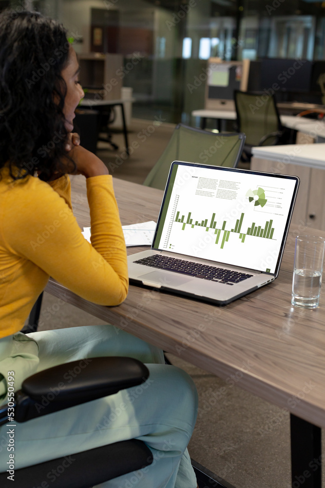 Poster vertical of biracial businesswoman using laptop in empty office with graph and data on screen