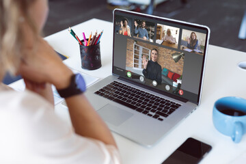 Caucasian businesswoman at desk making laptop video call with diverse colleagues waving
