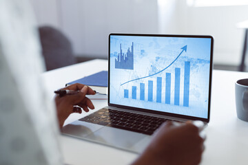 Hands of african american businesswoman at desk using laptop with blue graphs on screen