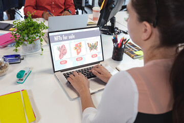 Caucasian businesswoman using laptop in office with nfts for sale on screen