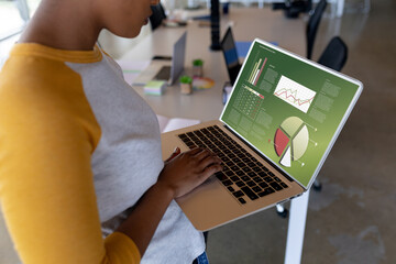 Midsection of african american woman using laptop in office with charts and data on screen