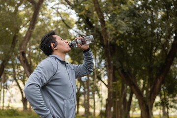 Fototapeta na wymiar Thirsty runner man is drinking water bottle with earphone after exercise cardio on path in forest background.