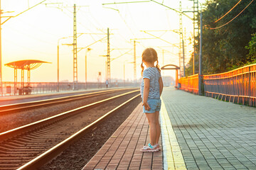 a little girl stands on the platform of the railway station in the evening sunset time and waits for the train