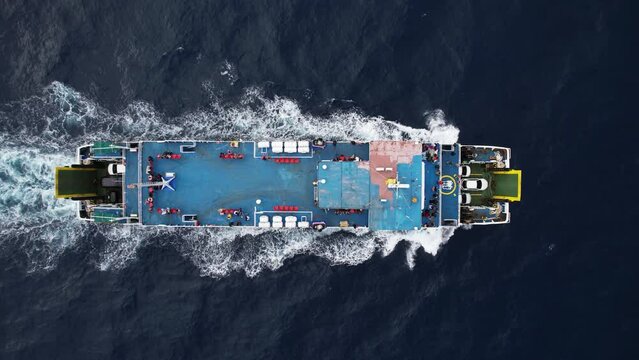 Top-down aerial trucking shot of sailing Ro-Ro passenger ship. Cars seen on vehicles deck, few people sit on empty upper deck. Small waves break to white splashes at stem of vessel