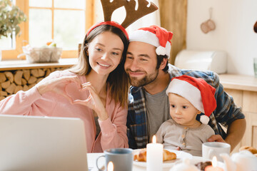 Happy loving young caucasian family of three, parents and toddler son daughter child kid celebrating Christmas New Year together, showing heart shape gesture on videocall on laptop