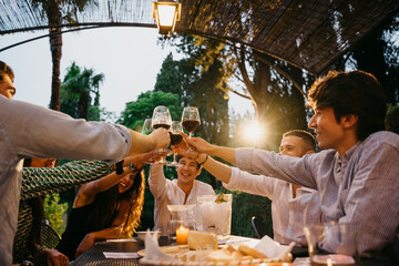 Group of friends outside eating and have fun together toasting with red wine glasses at sunset at hotel restaurant where are staying - Cheerful people laugh and joke evening at sunset - Party at home