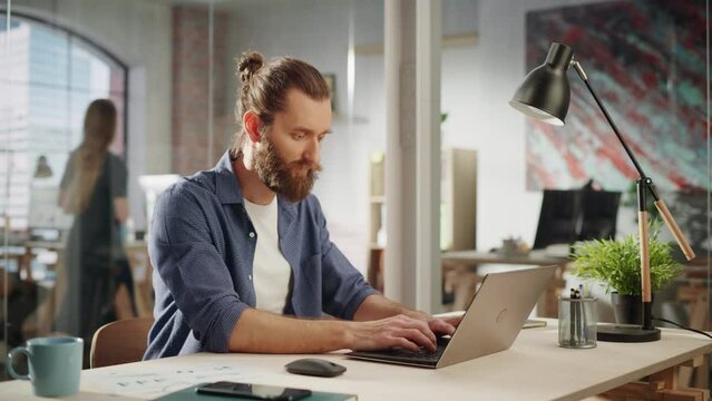 Handsome Long-Haired Bearded Manager Sitting at a Desk in Creative Office. Young Stylish Man Using Laptop Computer in Marketing Agency. Colleagues Working in the Background.