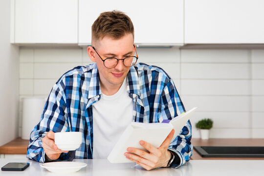 A man in a plaid shirt and glasses drinks coffee and reads a book in his kitchen