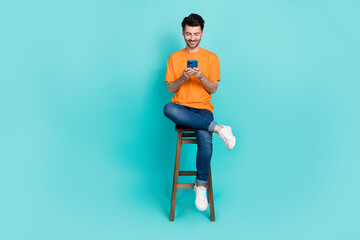 Full length photo of attractive man sit stool hold telephone device share news post repost typing space isolated on cyan color background