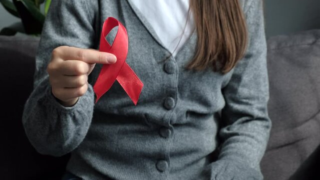 Close up of young woman sit on couch holding small red ribbon breast cancer symbol support people patient with illness, female volunteer keep AIDS HIV disease awareness, healthcare, medicine concept