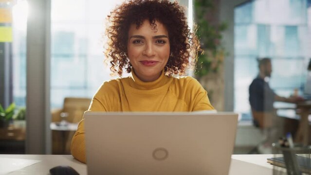 Portrait of an Attractive Arab Female Sitting at a Desk in Creative Agency. Young Stylish Manager with Curly Hair Smiling, Looking at Camera. Manager Working in Modern Company.