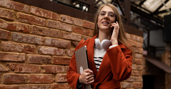 young woman rushing to work with a smile on her face with a laptop under her arm talking on a mobile phone against the backdrop of the city wall of the building