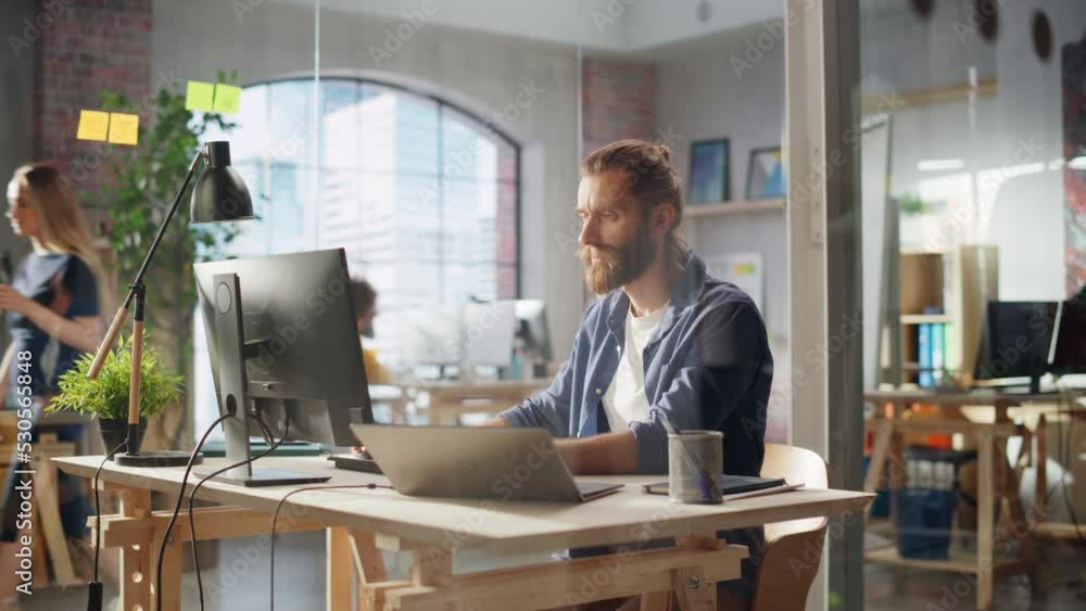 Poster handsome long-haired bearded manager sitting at a desk in creative office. young stylish man using l