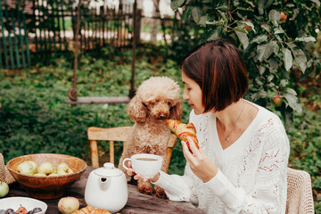 young beautiful asian woman sitting at table resting and drinking tea after harvesting in autumn in back yard with dog, fall and harvest, zero waste life, eco-friendly simple living, apples, croissant