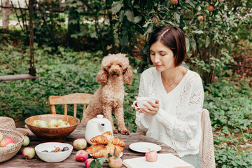 young beautiful asian woman sitting at table resting and drinking tea after harvesting in autumn in back yard with dog, fall and harvest, zero waste life, eco-friendly simple living, apples, croissant