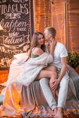 A young and attractive couple, a guy and a girl are sitting and hugging on a bed in a room decorated for Christmas with garlands. Christmas mood