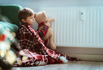 cute little boy wrapped id plaid sitting by heater warming teddy bear paws