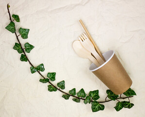 Fototapeta na wymiar Disposable bamboo cutlery in disposable paper cups on canvas background and natural ivy branch. Vertical image.