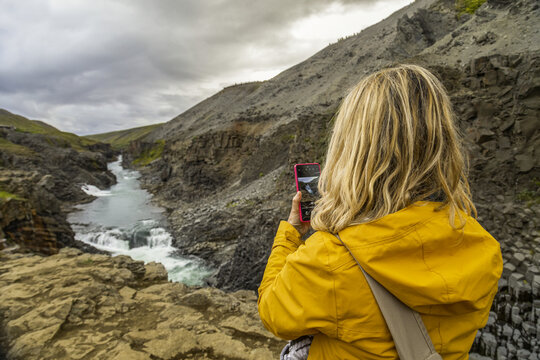 Donna bionda in viaggio mentre fotografa con un cellulare un canyon con un fiume in Islanda