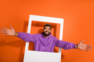 Photo of cheerful nice young man raise opened hands through paper album card isolated on orange color background