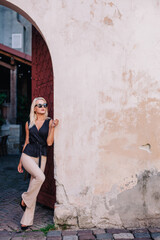 Fototapeta na wymiar a blonde beautiful girl in sunglasses, a black blouse and beige pants poses against the background of an old collar
