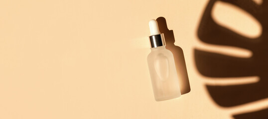 Frozen glass dropper bottle with white cap on beige background with tropical leave shadow. Pipette...