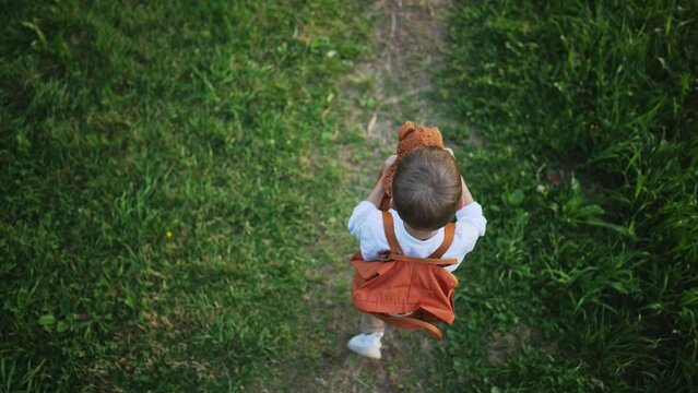 Top view of baby boy walking along path and holding toy bear in hands. Child in school uniform with backpack. Preparation and back to school. Walking on road in nature outdoors in forest or park.