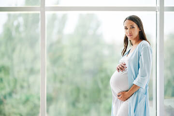 Portrait of young pregnant woman standing near window holding hands on her belly at modern home