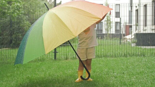 Little girl in yellow rubber boots under a large colorful umbrella in the midst of summer rain.