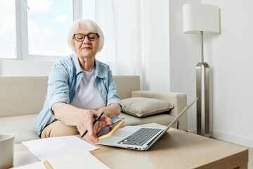 an elderly woman is sitting at home on the couch at work with a laptop on the table and notebooks for notes picking up a smartphone