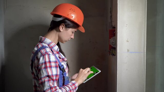 electrical engineer woman is drawing electrical panel diagram by tablet, looking inside electric board, repair and renovation in industrial building. Construction, electricity and technology