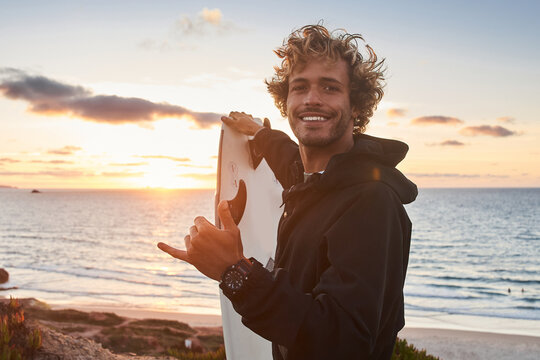 Surfer man standing on the hill with surfboard and posing for camera with shaka gesture