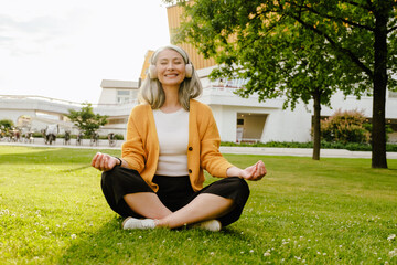 Grey asian woman in headphones meditating while sitting on grass