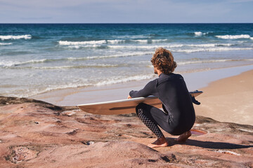 Surfer kneeling towards the ocean with his board at the hands and looking at the distance